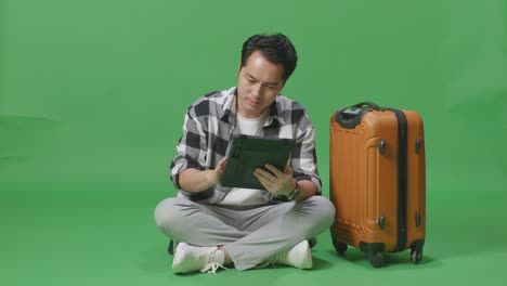 full body of asian male traveler with luggage using a tablet while sitting in the green screen background studio