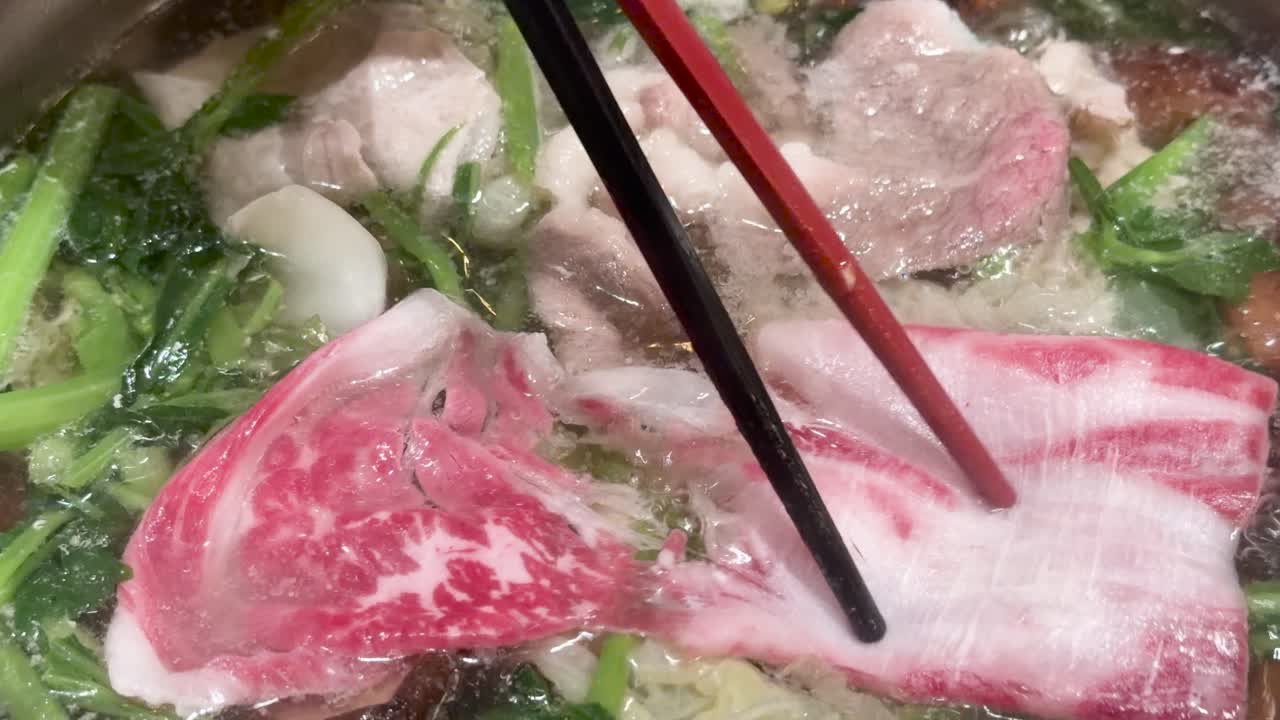 Chopsticks add marbled beef to simmering hotpot with vegetables, mushrooms, and steaming broth, overhead view
