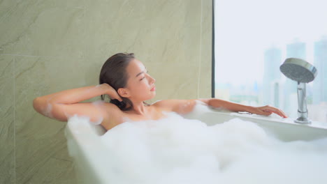 a beautiful young woman lounges in a bubble bath as she takes in the modern world through her window from her high-rise building