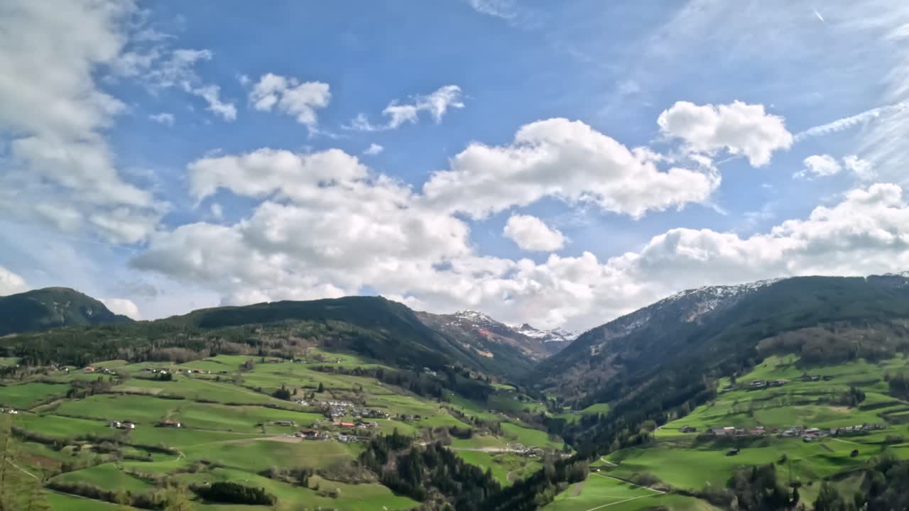 Aerial-style view of a lush green valley surrounded by alpine mountains under a partly cloudy sky, captured on the way to Innsbruck, Austria