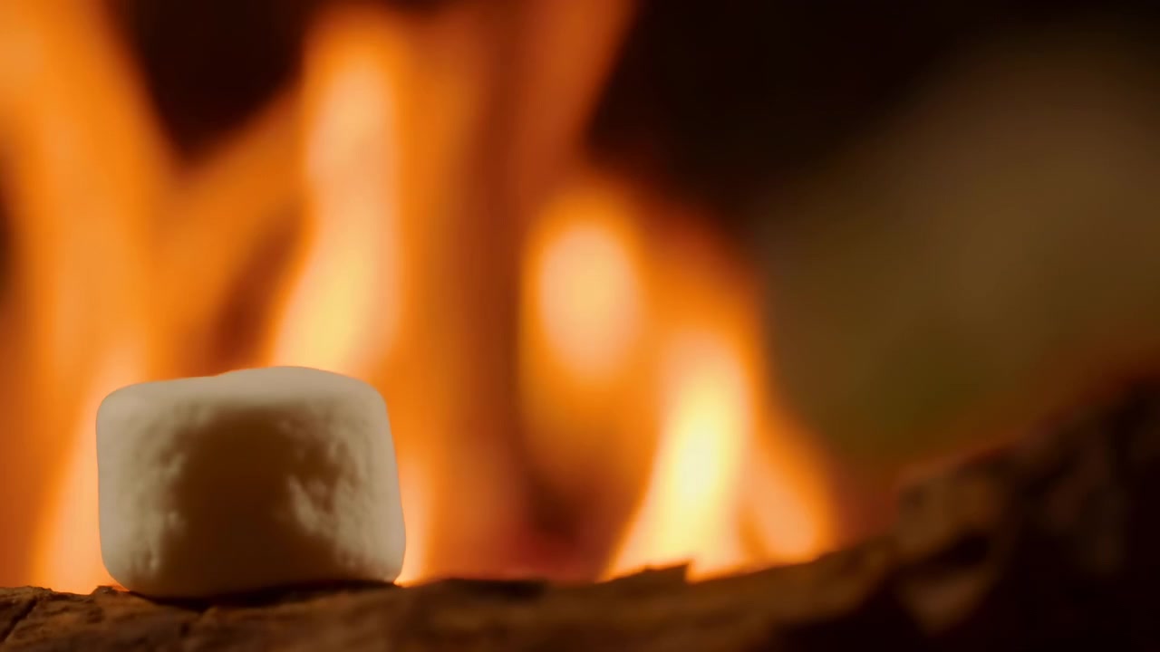 Un primo piano ravvicinato mostra fiamme arancioni brillanti che si alzano sopra braci ardenti e legno carbonizzato, con uno sfondo scuro e sfocato e una luce bassa e calda che crea un