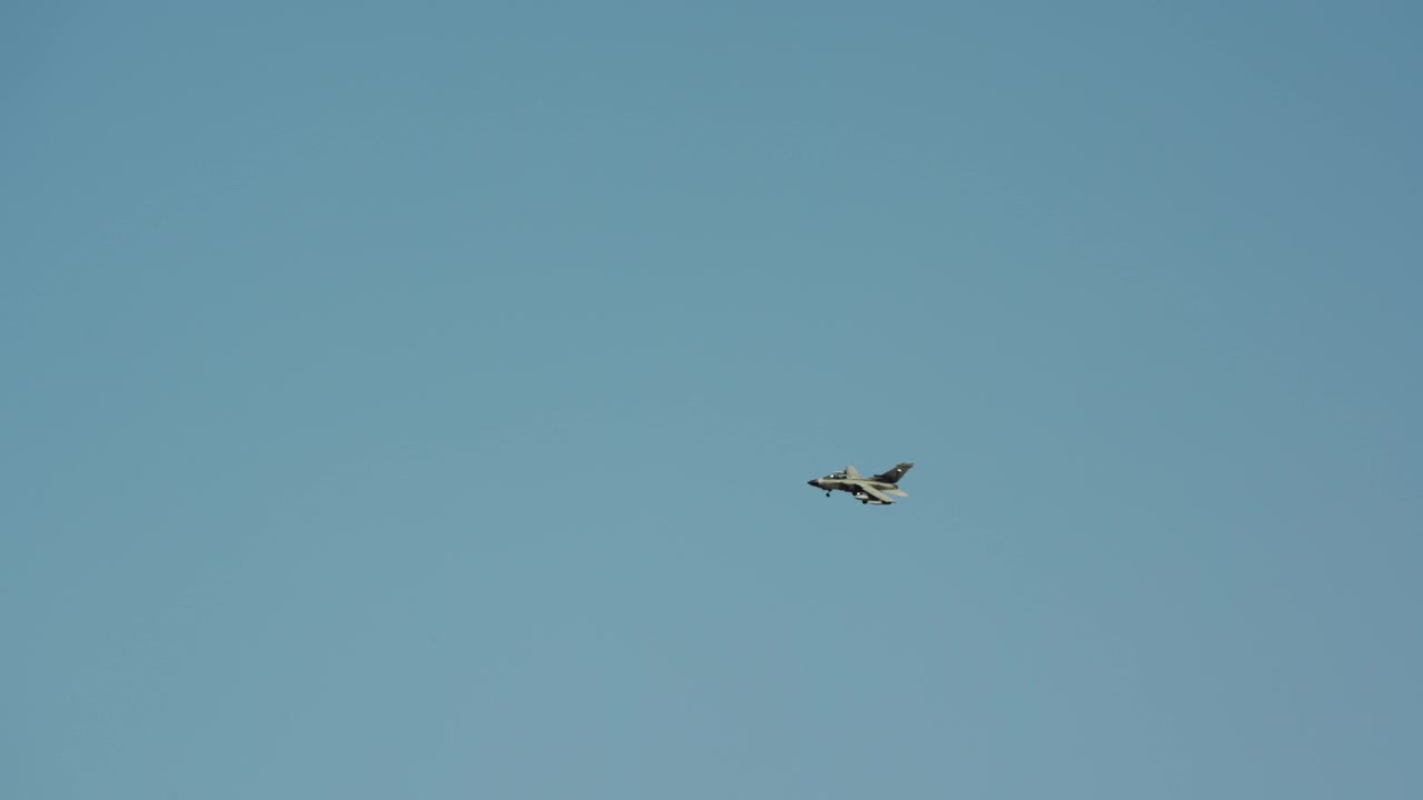 A military fighter jet flies across a clear blue sky in slow motion, maintaining its altitude during a daytime flight