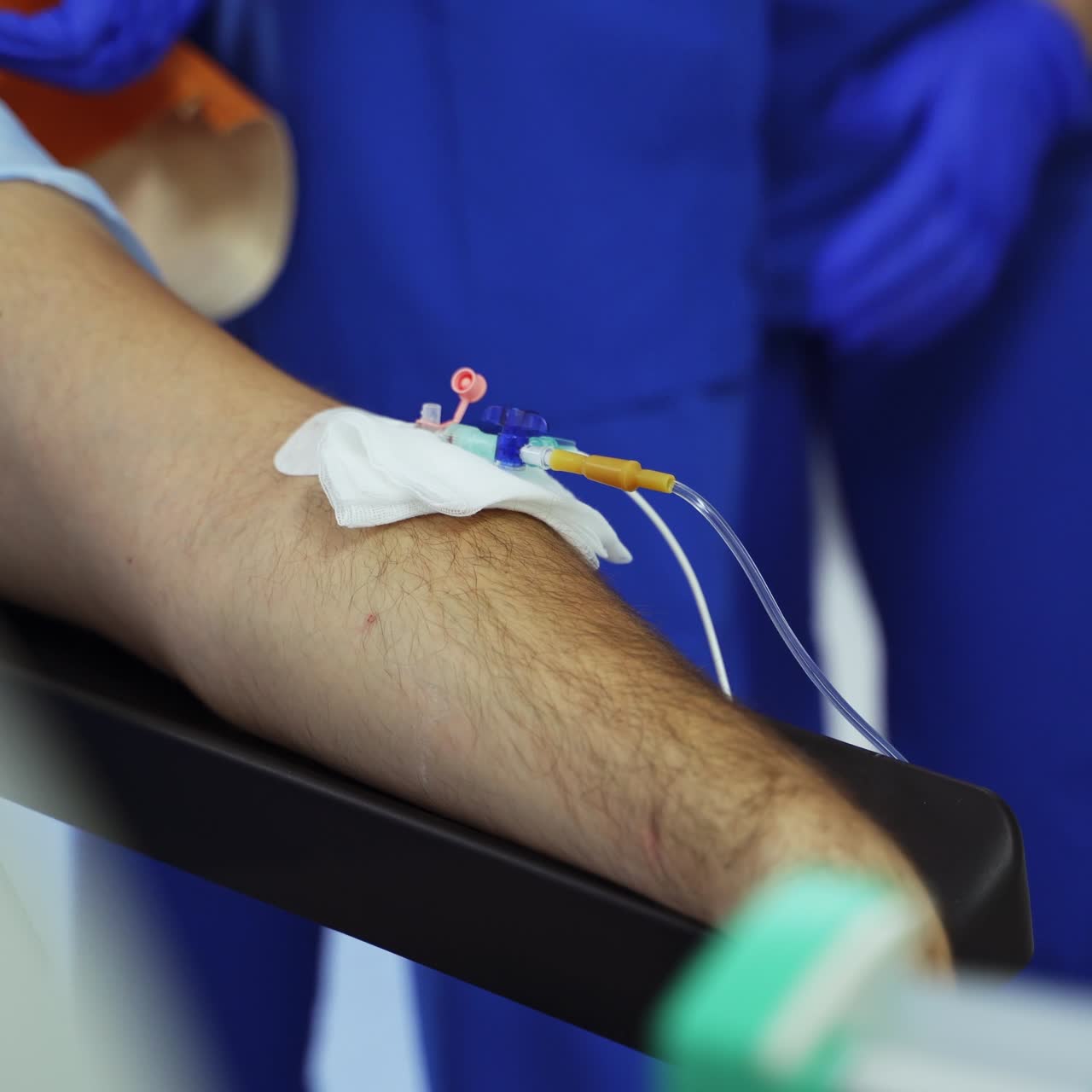 Patient's male hand with attached catheter and tubes of drop counter. Medical staff walks around preparing the patient for operation