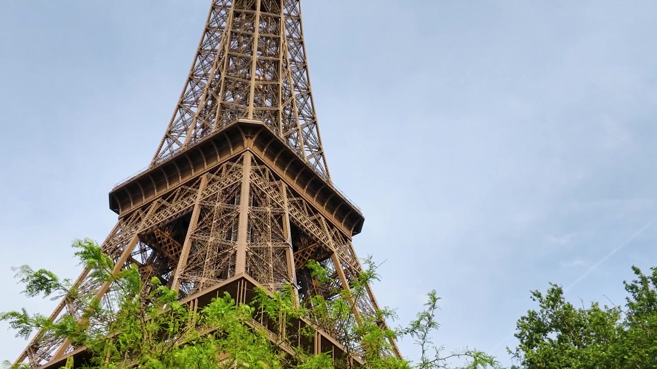 Un'inquadratura statica dal basso cattura l'iconica torre in traliccio di ferro dietro alberi verdi e rigogliosi.