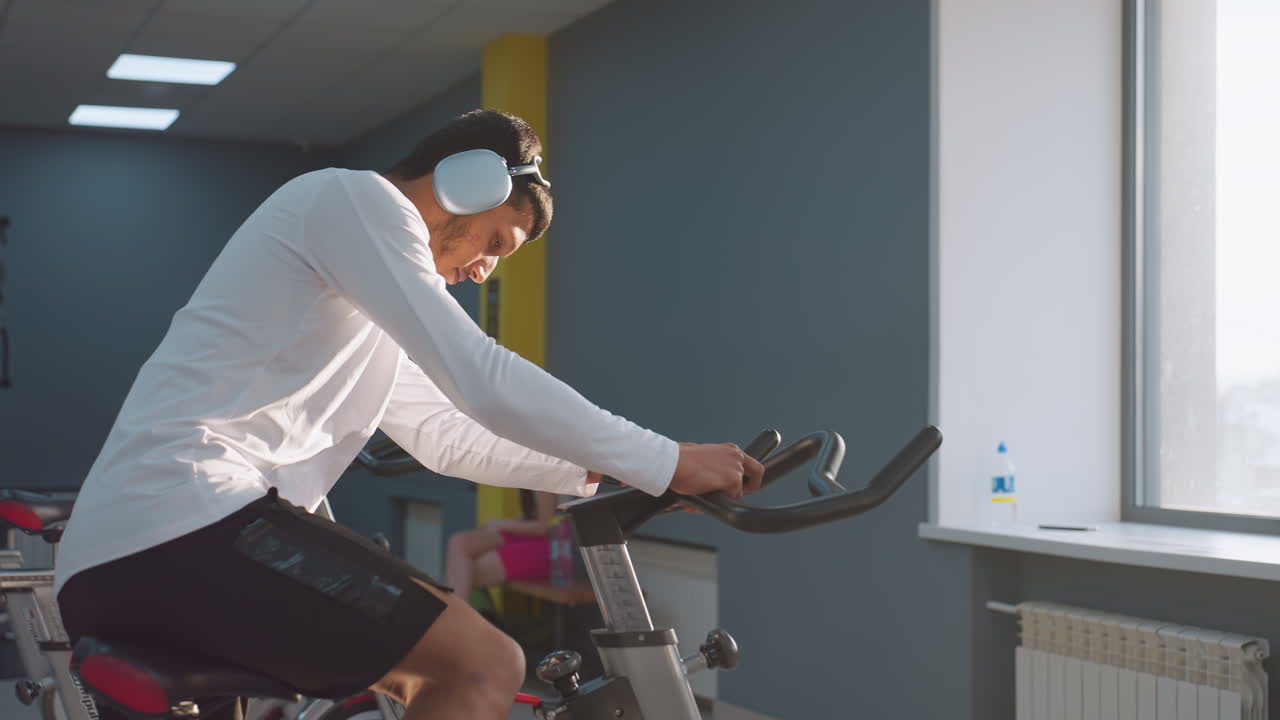 Athlete on headset mounts stationary bike and begins workout under natural light streaming through gym windows, with water bottle on sill and other people exercising in soft background