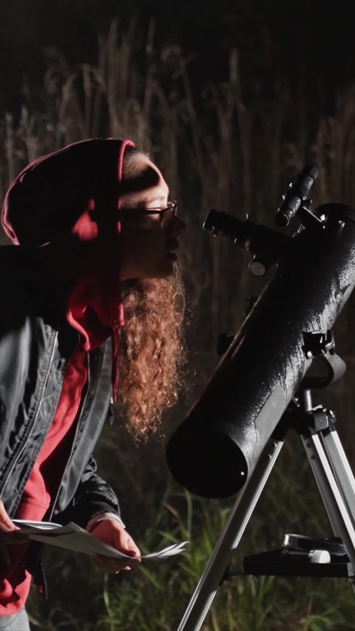 Individual writes and looks at notes by tripod mounted. She adjusts papers while looking down during night time. Dark outdoor area has visible plants behind woman engaged