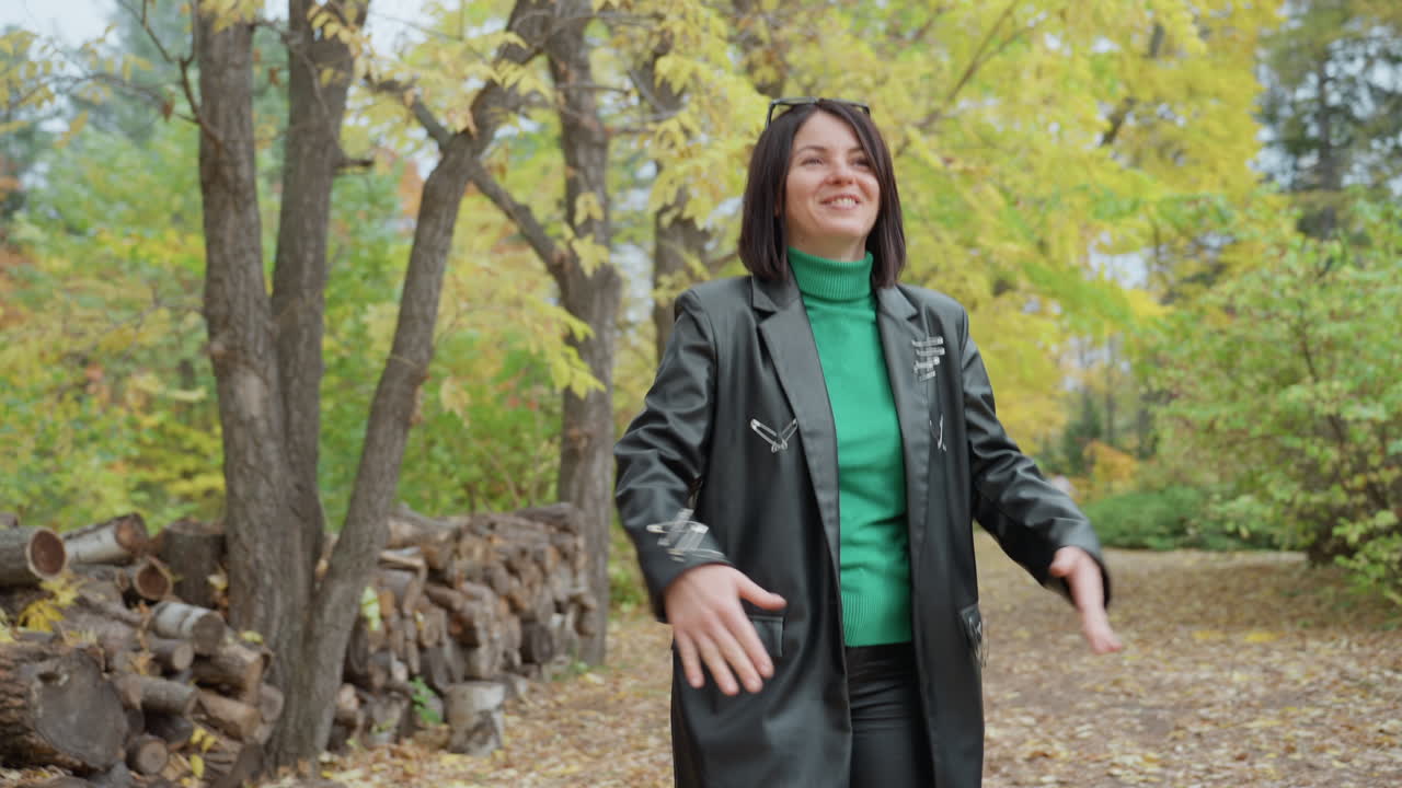 Woman in leather jacket laughing on forest path with stacked logs, green turtleneck visible, hands clasped, candid smile, falling leaves, calm breeze, moody rock style, casual fashion portrait evoking
