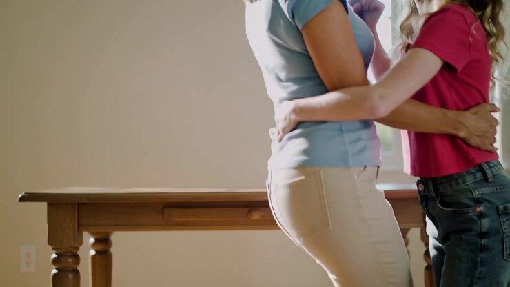 Hugging mother, teen daughter swaying in lit nook by wooden table, window, light-blue and pink tops