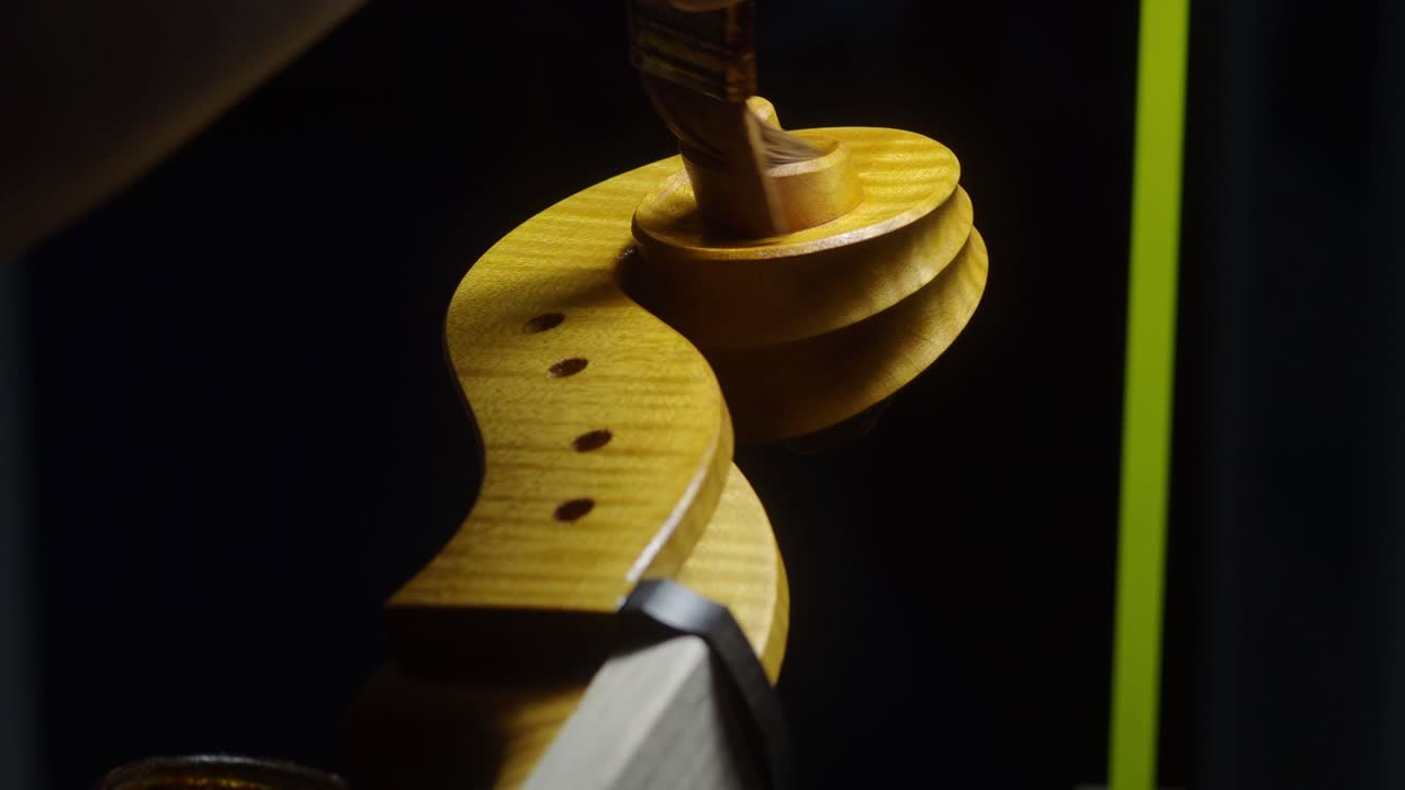 Skilled woodworker brushing protective varnish onto wooden cello scroll and pegbox surface, highlighting meticulous handcrafting technique in traditional instrument making workshop, real time
