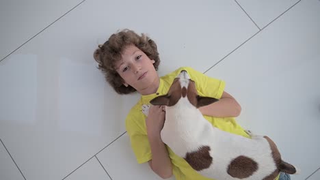 top view of a blond boy with curly hair lying on the floor and caresses his dog