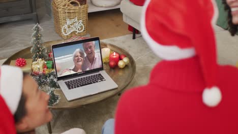 Diverse-family-with-santa-hats-using-laptop-for-christmas-video-call-with-happy-couple-on-screen