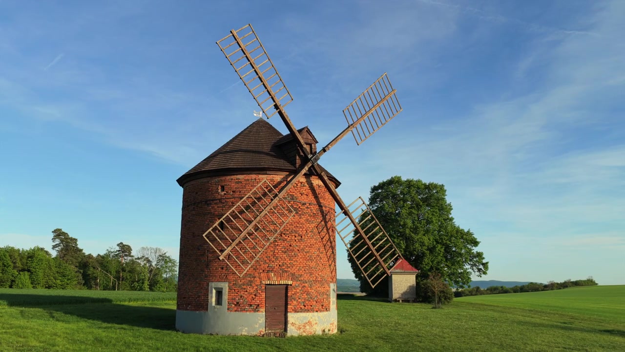 Vue aérienne par drone d'un moulin à vent historique en briques, solitaire, avec quatre ailes sur une colline verdoyante sous un vaste ciel bleu avec des terres agricoles vallonnées en arrière-plan, Moravie, République tchèque.