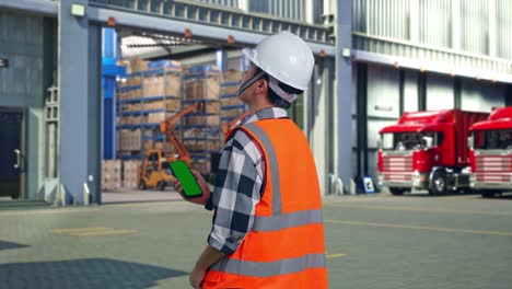 back view of asian male engineer with safety helmet working on a green screen smartphone and looking around while standing , outside of logistics distributions warehouse