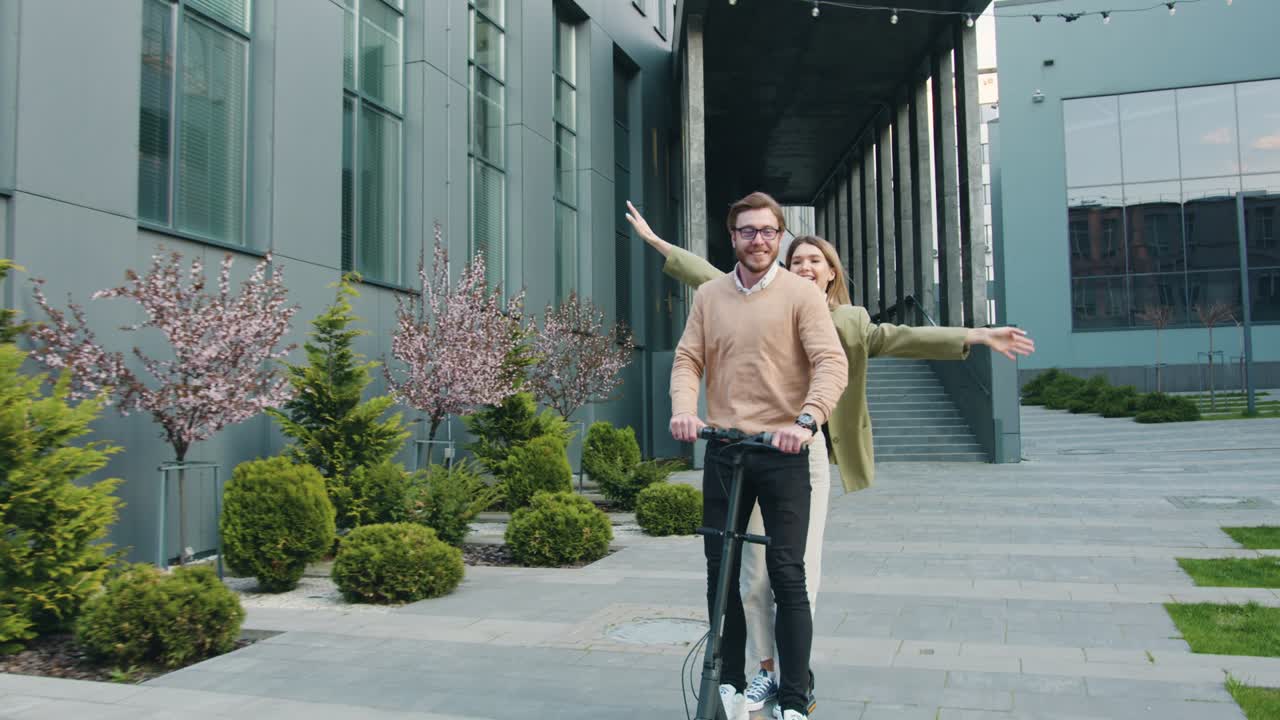 Happy young caucasian couple smiling and enjoying riding of electric scooter together in city near modern office centre. Outside, lifestyle concept