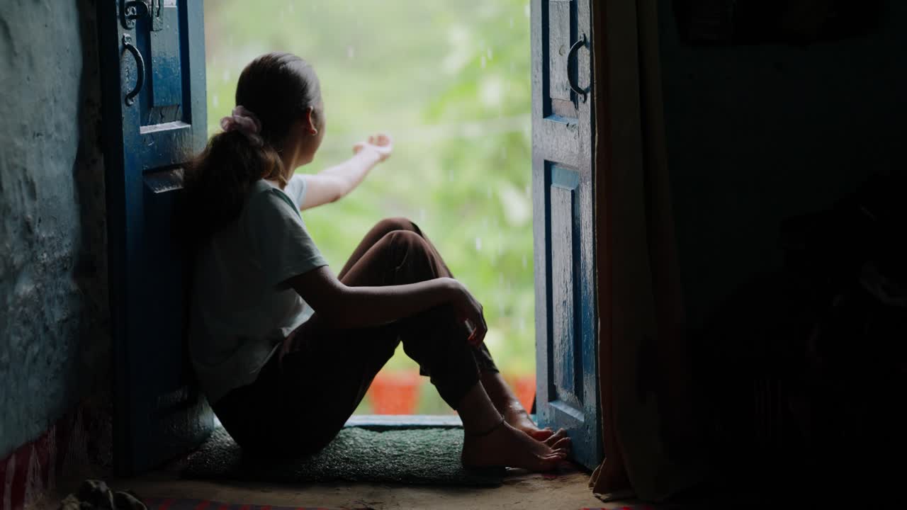 Village girl sitting quietly at her front door, smiling as rain pours outside, cinematic countryside scene, 4k video