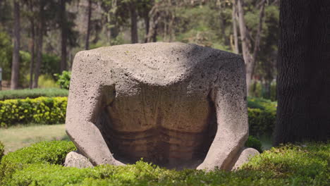 headless olmec statue sits facing camera under dappled sunlight in wooded landscape, parque hundido mexico city