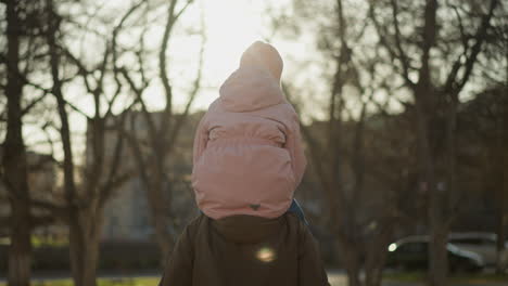 back view of a little girl in a pink cap and jacket sitting on her dad's neck as they walk through a park. the sun shines through the trees, creating a warm, serene atmosphere