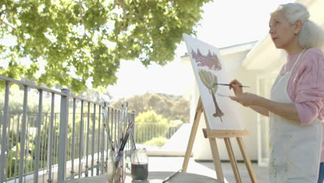 happy senior biracial woman painting at balcony at home, slow motion