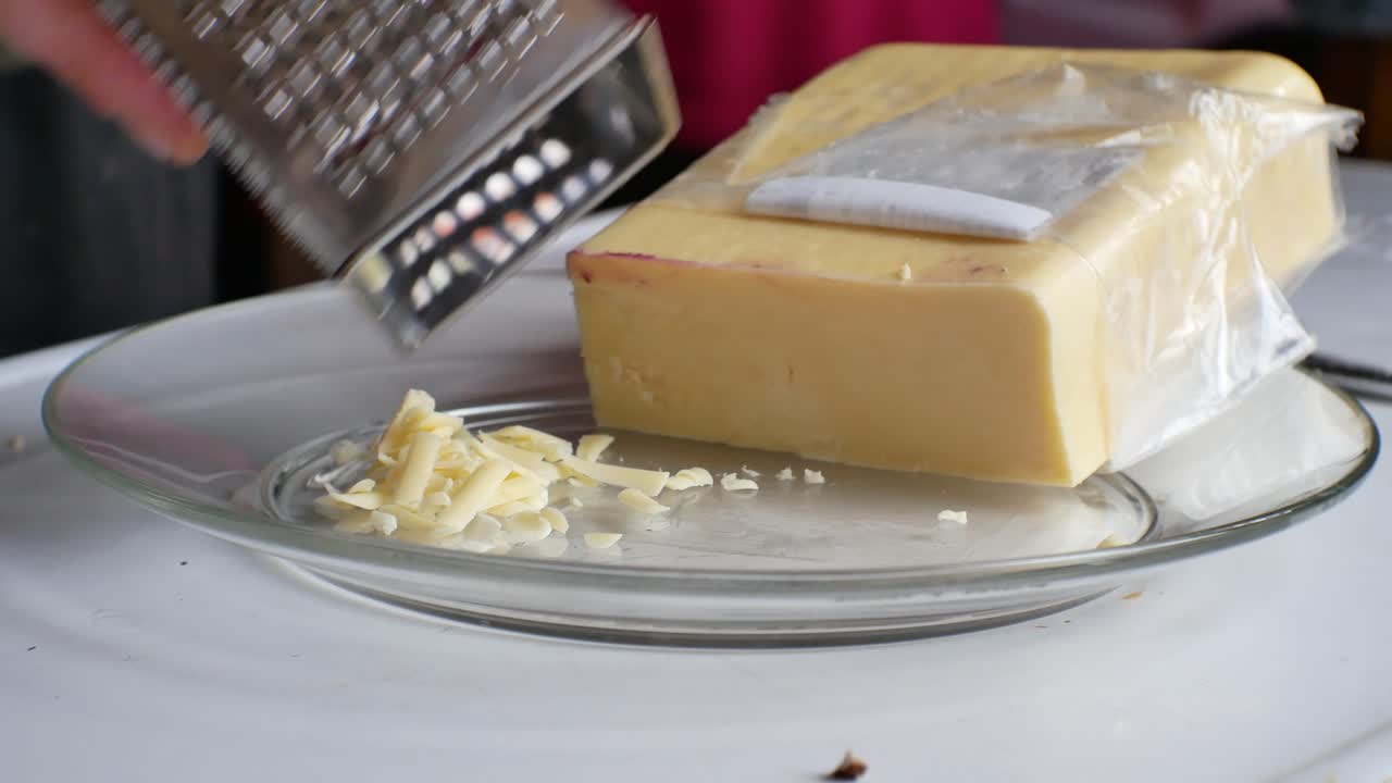 cierre las manos masculinas recogiendo un bloque de queso cheddar amarillo en rodajas - rallándolo en una pila en un pequeño plato de vidrio en la cocina de la casa