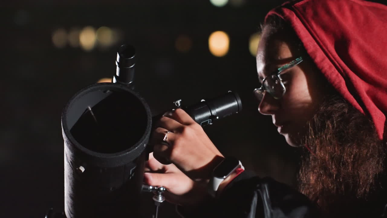 Hvidskægget astronom justerer teleskop, by, lys bokeh i baggrunden, rød hættetrøje og briller, nærbillede af hænder der justerer okular, fokuseret udtryk, observationsvidenskab under stjernerne