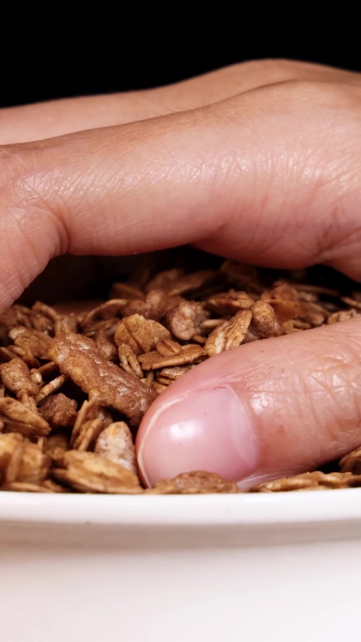 Una mano mescola la granola in una ciotola bianca, creando un movimento dinamico su uno sfondo nero con inquadrature ravvicinate.