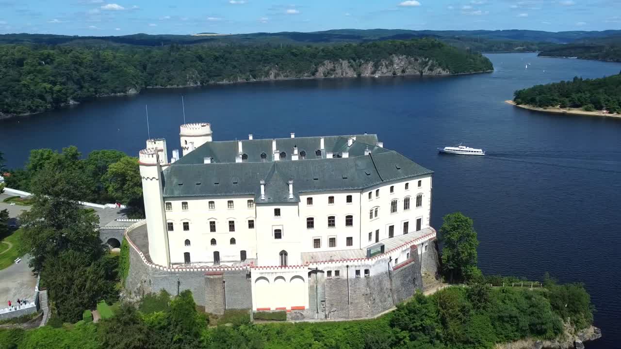 Orlik Castle in Czech Republic, nestled beside water with scenic hills under bright sky, aerial establishing orbit