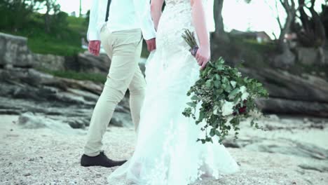 bride and groom walking on beach, bride has flowers in her hand - slow motion