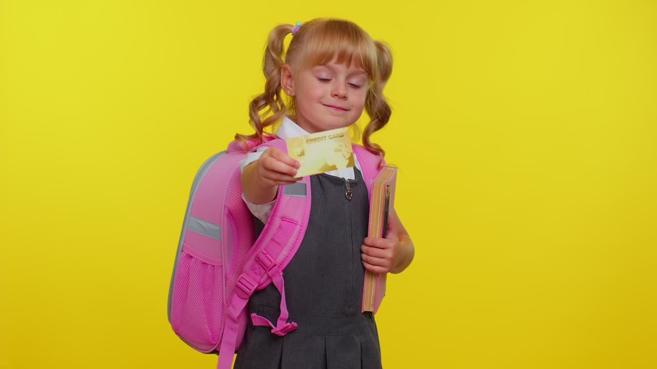 Bambina studentessa allegra e divertente in uniforme che tiene in mano una carta di credito bancaria e mostra il pollice in su.