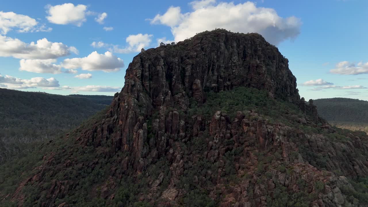 Drone camera circles Timor Rock, revealing rugged volcanic formation, dry forest, and open plains under bright daylight with scattered clouds and natural lighting