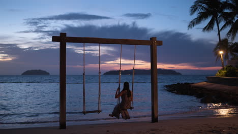 Mujer-Asiática-Solitaria-Columpiándose-Sola-En-Un-Columpio-De-Cuerda-En-La-Playa-Para-Parejas-En-La-Pintoresca-Y-Espectacular-Puesta-De-Sol-Con-Siluetas-De-Islas-Y-Esponjosas-Nubes-Moradas---Vista-Trasera