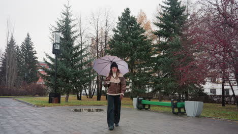 young girl holding umbrella wearing black knit cap, brown shearling jacket, and black trouser walking calmly through paved park on cold overcast day, light snow falling, surrounded by evergreens