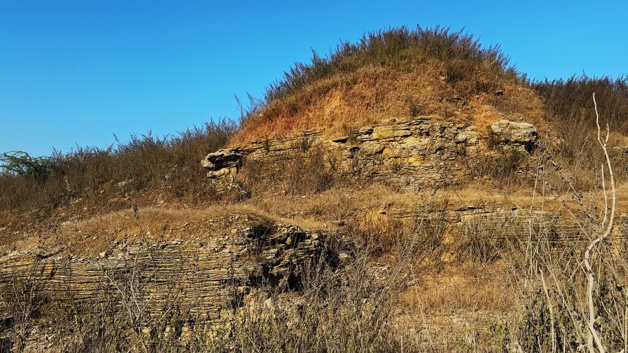 La camera in orbita rivela una collina rocciosa stratificata con erbe secche e arbusti, le cui texture in pietra esposta sono erose sotto un cielo azzurro, catturando il terreno aspro e i modelli di erosione