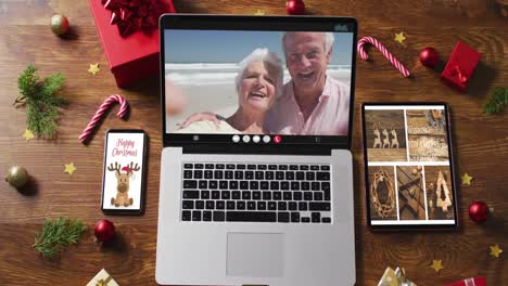 Caucasian-senior-couple-waving-on-video-call-on-laptop,-with-smartphone,-tablet-and-decorations