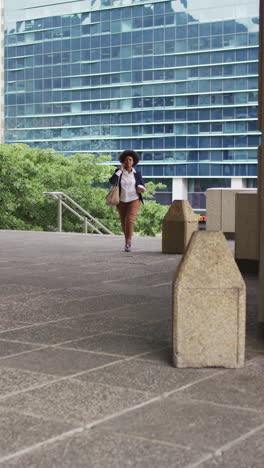 video vertical de una feliz mujer afroamericana caminando por la ciudad con café, hablando en su teléfono inteligente