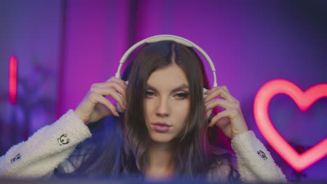 overhead view young woman at gaming desk placing white headphones on head while resting hand on desk under neon heart pink purple lighting with dual monitors rgb keyboard glow for live stream setup