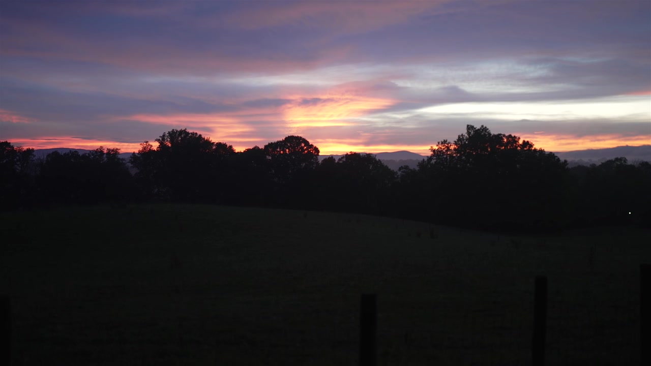 Enchanting panoramic footage of a rural sunset, sweeping from left to right. The dreamy scene captures the twilight sky's soft hues above a serene countryside, with silhouetted trees framing horizon.