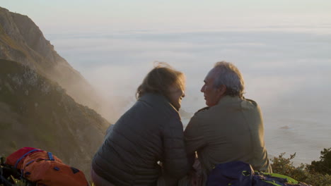 vista trasera de una pareja mayor sentada en la cima de la montaña y besándose mientras disfruta de la hermosa vista