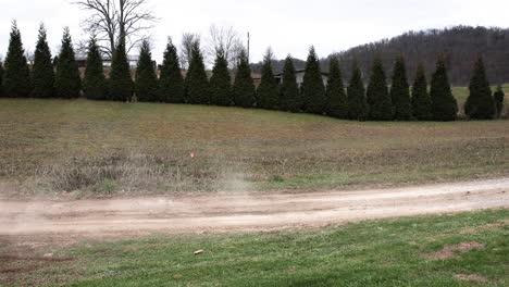 Weißer-Rallye-Geländewagen,-Der-Mit-Einer-Rosa-Flagge-Im-Hintergrund-Durch-Einen-Schlammigen-Feldweg-Rast-Und-Eine-Staub--Und-Schmutzspur-In-Der-Luft-Hinterlässt,-Während-Er-Mit-Einer-Reihe-Von-Evergreens-Im-Hintergrund-Vorbeirast