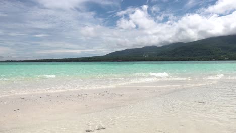 Clear-turquoise-blue-ocean-waves-washing-ashore-on-a-white-sandy-tropical-island-beach-with-lush-green-trees-covering-volcanos-and-mountains-in-the-background