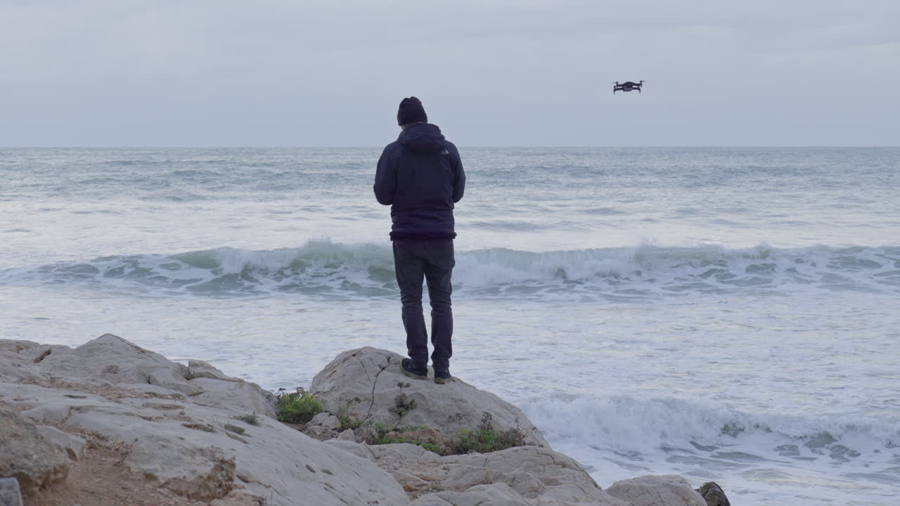 무인 항공기가 날아갈 때 남자는 폭풍우가 치는 바다를 바라보며 절벽에 서 있습니다.
