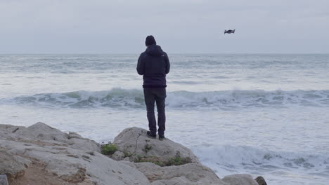 El-Hombre-Se-Para-En-Un-Acantilado-Mirando-Hacia-El-Mar-Tormentoso,-Mientras-El-Dron-Vuela