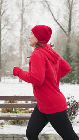 side view of tired lady jogging along snowy park pathway in red hoodie and black leggings surrounded by benches colorful bushes frosty winter trees and distant snow dusted offices