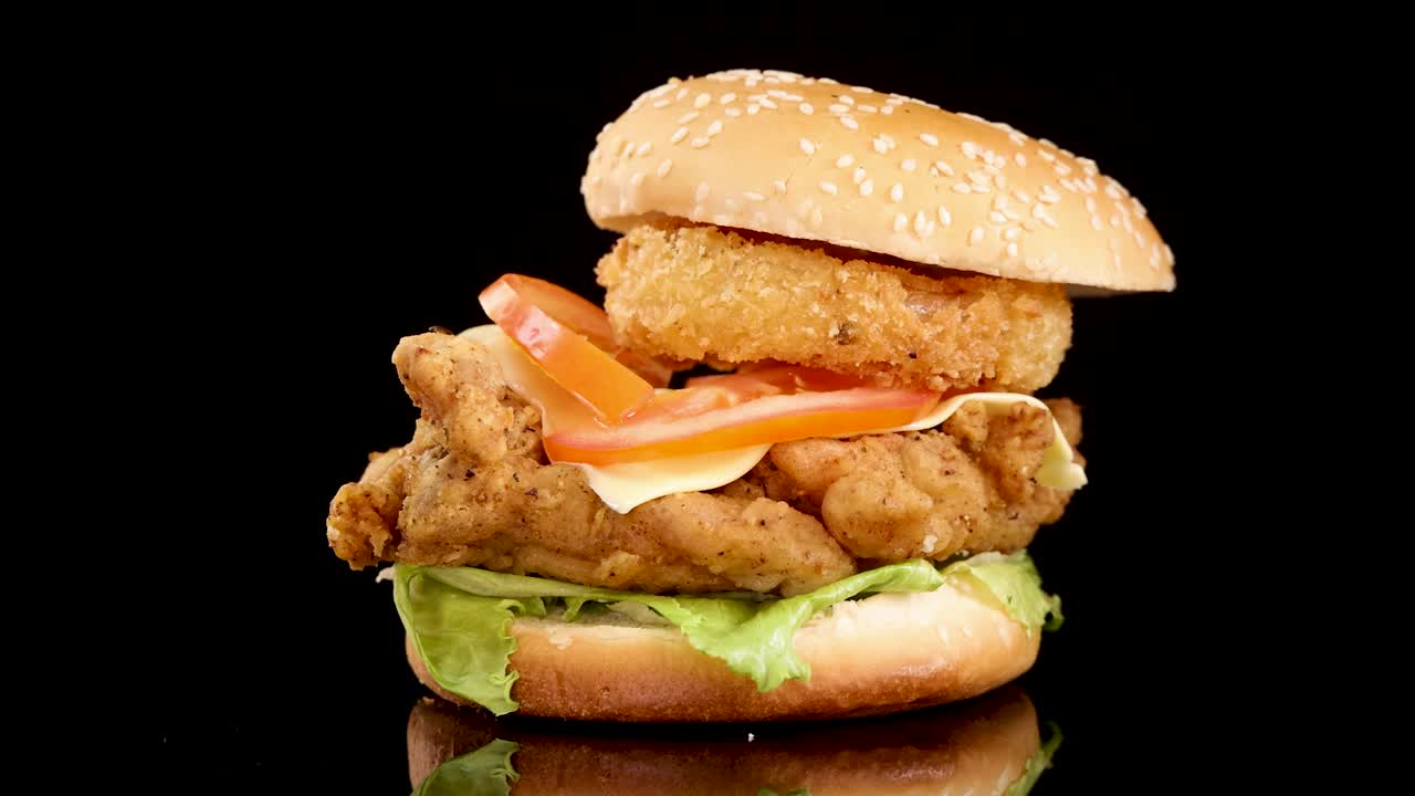 Un panino ai semi di sesamo cade su un hamburger di pollo fritto impilato con lattuga, pomodoro, formaggio e anello di cipolla, su uno sfondo nero con un'illuminazione da studio drammatica.