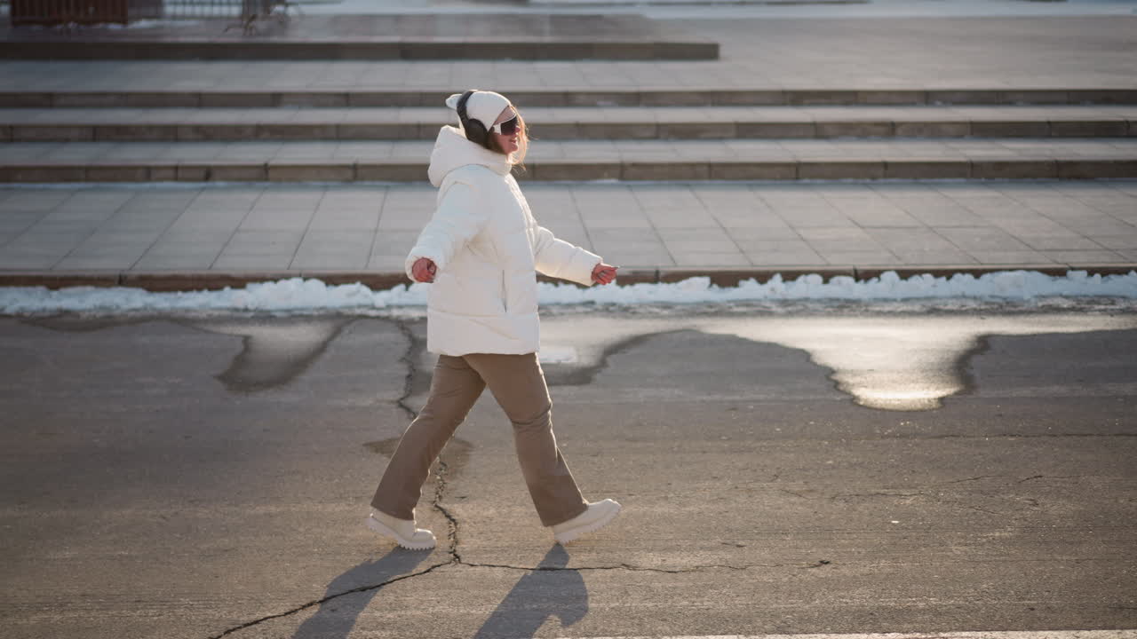 Young artist walking down tiled street wearing headphones and beanie, moving to music beat against snowy curb and urban stair backdrop under winter sunlight, expressive arms and joyful smile
