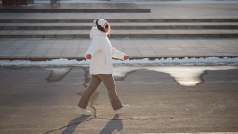 young artist walking down tiled street wearing headphones and beanie, moving to music beat against snowy curb and urban stair backdrop under winter sunlight, expressive arms and joyful smile