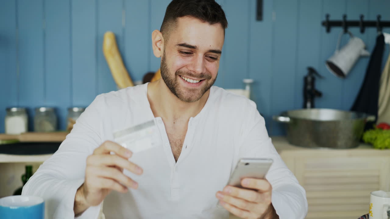 Man using mobile phone and credit card for online payment in kitchen.