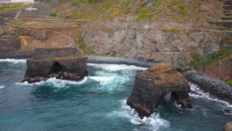 Los-Roques-Beach,-Tenerife:-aerial-view-in-orbit-of-the-two-rock-formations-and-the-crashing-sea