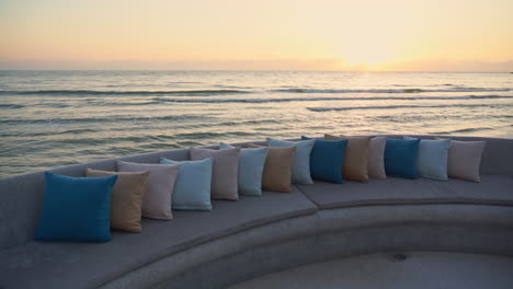 ocean waves roll toward the shore in the background, foreground an empty padded sitting area covered end to end with colorful pillows