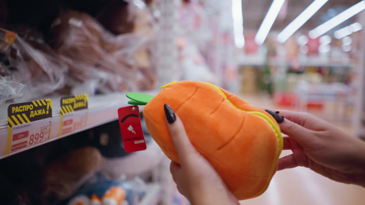 제품 세부 사항을 관찰하는 동안 상점 선반에 세련된 오렌지색 지갑을 잡는 여성의 뒷면 클로즈업, 다양한 액세서리와 프로모션 판매 표지판을 가진 소매 통로를 탐색하는 구매자