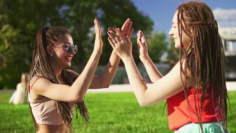 happy women with dreads sitting on grass in summer park. young friends talking and playing outdoors having fun. mulatto woman